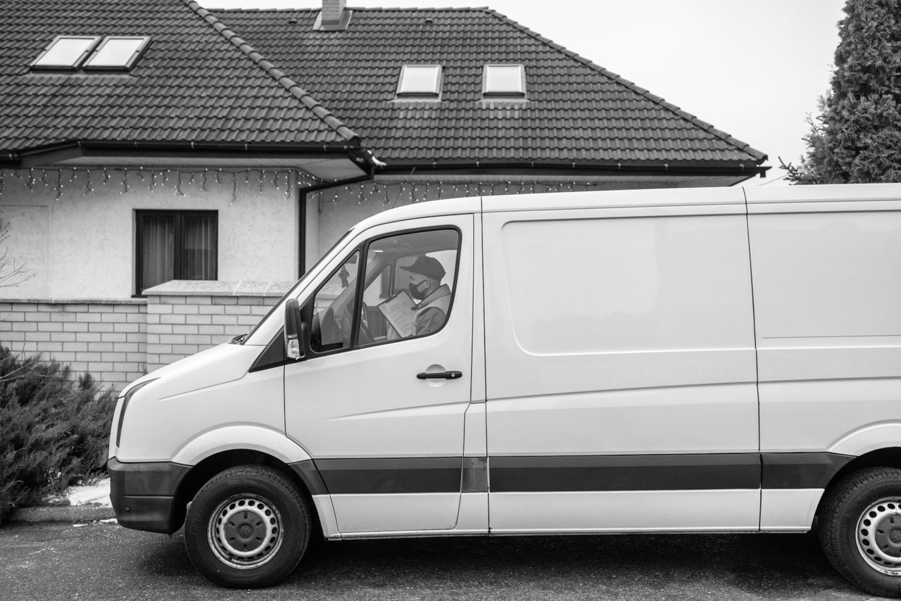 services-03 Side view of a white delivery van parked outside a residential house with driver reading a document.