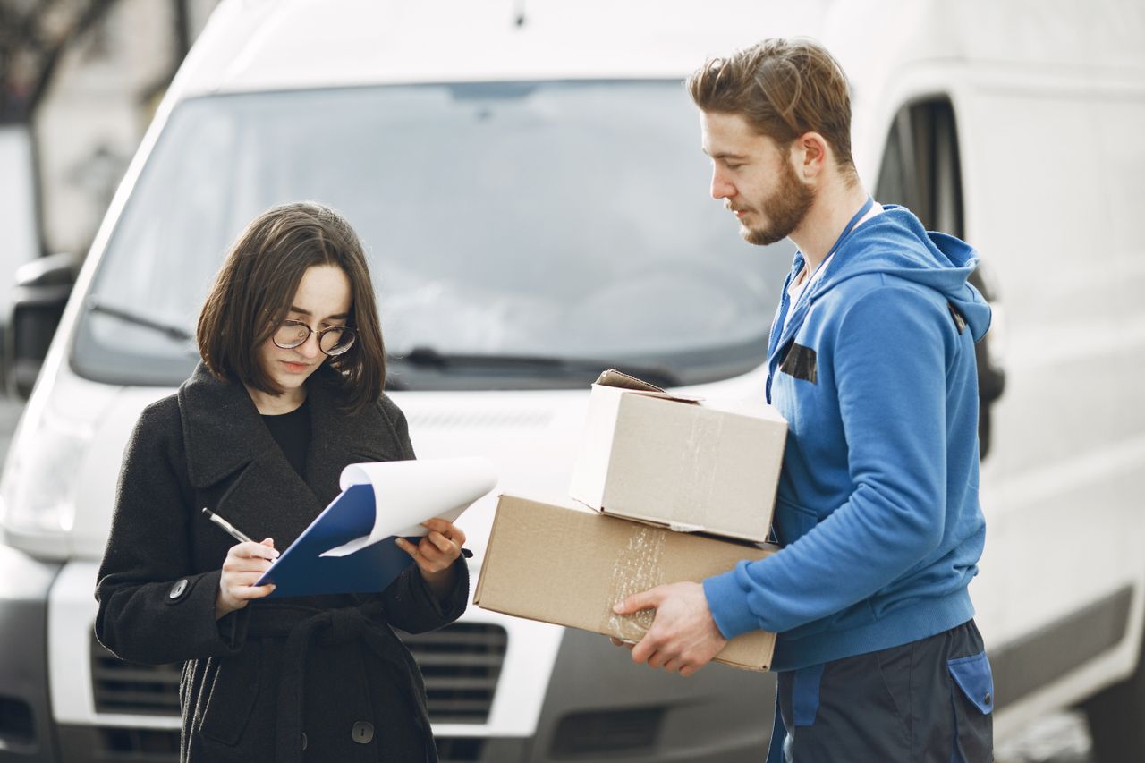 who-we-are A courier in blue hands packages to a woman signing documents in front of a delivery van.